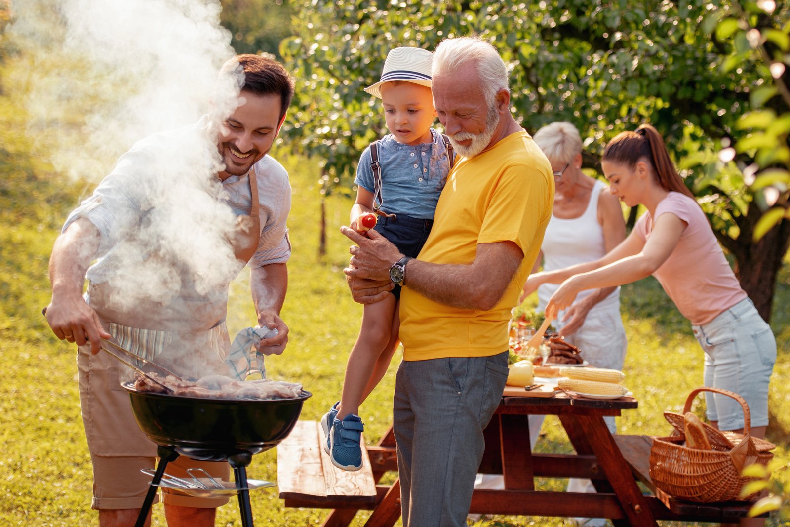 Grandfather holding his grandson at a backyard barbecue, with family gathered around the smoking grill